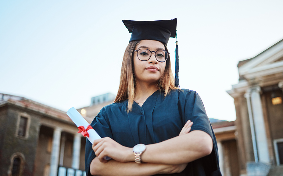 Young woman in graduation gown and cap proudly holds her diploma in her hand.