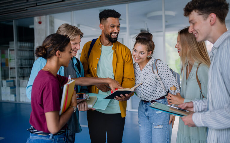 A group of young adults stand together, laugh and look at an open book.