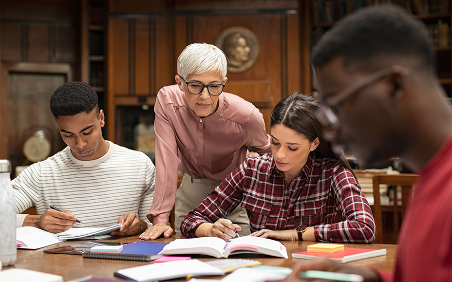An elderly woman supports three students in their studies in a library.